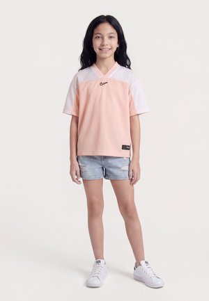 Κορίτσι όρθιο, φοράει αθλητικό πουκάμισο Nike σε ροδακινί και λευκό, σκισμένο τζιν σορτς και λευκά αθλητικά παπούτσια, χαμογελώντας με τα χέρια χαλαρά στο πλάι.