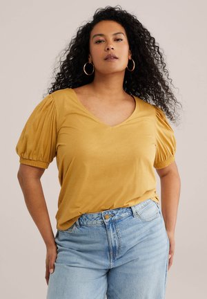 Kvinde med krøllet hår iført guld øreringe i hoops, en sennepsgul bluse med pufærmer og lyseblå cowboybukser med høj talje, stående med hænderne på hoften.