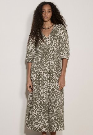 Vrouw met lang krullend haar, die een midi-jurk met V-hals draagt met groene en witte abstracte patronen en opgepofte driekwartmouwen.