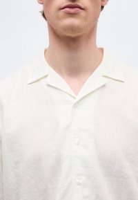 Νεαρός άνδρας που φοράει λευκό λινό πουκάμισο με κουμπιά και ανοιχτό γιακά, εμφανιζόμενος από τον λαιμό έως το μέσο του στήθους σε απλό φόντο.