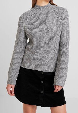 Nainen, jolla on harmaa ribbineulottu villapaita ja musta hame, jossa on napitus edessä, seisoo vaaleaa yksinkertaista taustaa vasten.