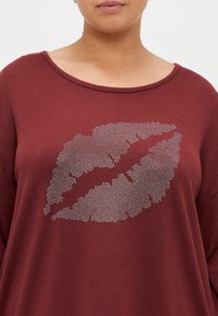 Sweater oversized em cor bordeaux com um gráfico de lábios em strass prateado no centro. Textura suave com um decote amplo e mangas longas.
