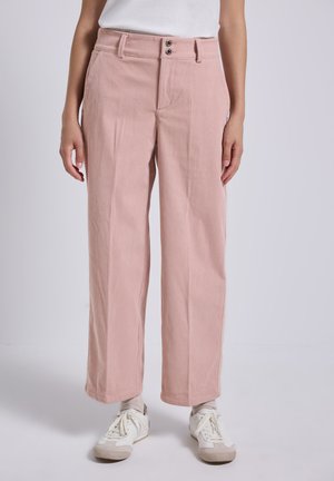 Vrouw in hoge taille, wijdvallende roze corduroy broek met witte sneakers en lichtgekleurde sokken op een effen achtergrond.