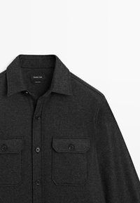 Camicia button-up in lana grigia scura con colletto e due tasche frontali a patta, ciascuna fissata con un bottone.