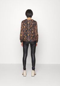 Blouse transparente noire à fleurs avec des manches longues et des poignets élastiques, associée à des leggings noirs brillants et des chaussures à talons blocs crème.