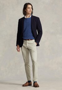 Mann iført marineblå blazer, blå kabelstrikket genser, beige bukser og brune loafers, stående mot en nøytral bakgrunn.