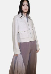 Kvinna iklädd en ljusbeige ulljacka med stora framfickor, mauve vida byxor, hållande en matchande mauve scarf i ena handen.