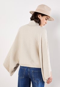 Maglione beige a maglia con coste verticali, colletto alto e maniche ampie. Abbinato a jeans blu, che mostrano una silhouette rilassata.