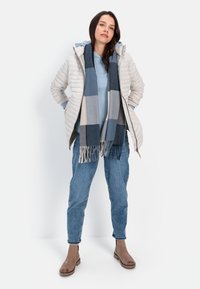 Lichtgrijze gewatteerde jas, blauwe hoodie, geruite sjaal in marineblauw en beige, blauwe jeans en bruine enkellaarzen met een gestructureerd oppervlak.
