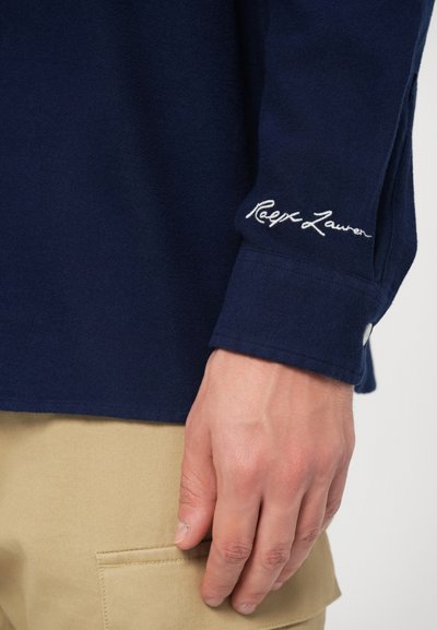 Polo din material moale, cu mânecă lungă, de culoare bleumarin, cu manșete cu nasturi. Prezintă un logo brodat alb "Ralph Lauren" în zona gulerului.