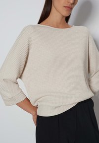 Hellbeiges, strukturiertes Sweatshirt mit lockerer Passform, weitem Halsausschnitt und 3/4-Ärmeln, kombiniert mit schwarzen Hosen, vor einem neutralen Hintergrund präsentiert.