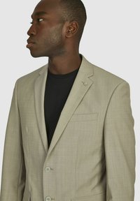 Lichtbeige blazer van gestructureerde stof, met een notch lapel, twee knopen en een enkele borstzak, gedragen over een zwart shirt.