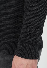 Svart stickad tröja med ett strukturerat mönster, med ribbade muddar och en mjuk kropp. Materialet verkar mjukt och elastiskt.