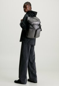 Zaino in nylon nero con apertura arrotolata, due tasche frontali e testo bianco. Il modello indossa abbigliamento scuro e si trova di fronte a uno sfondo neutro.