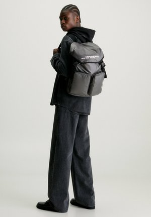 Zaino in nylon nero con apertura arrotolata, due tasche frontali e testo bianco. Il modello indossa abbigliamento scuro e si trova di fronte a uno sfondo neutro.