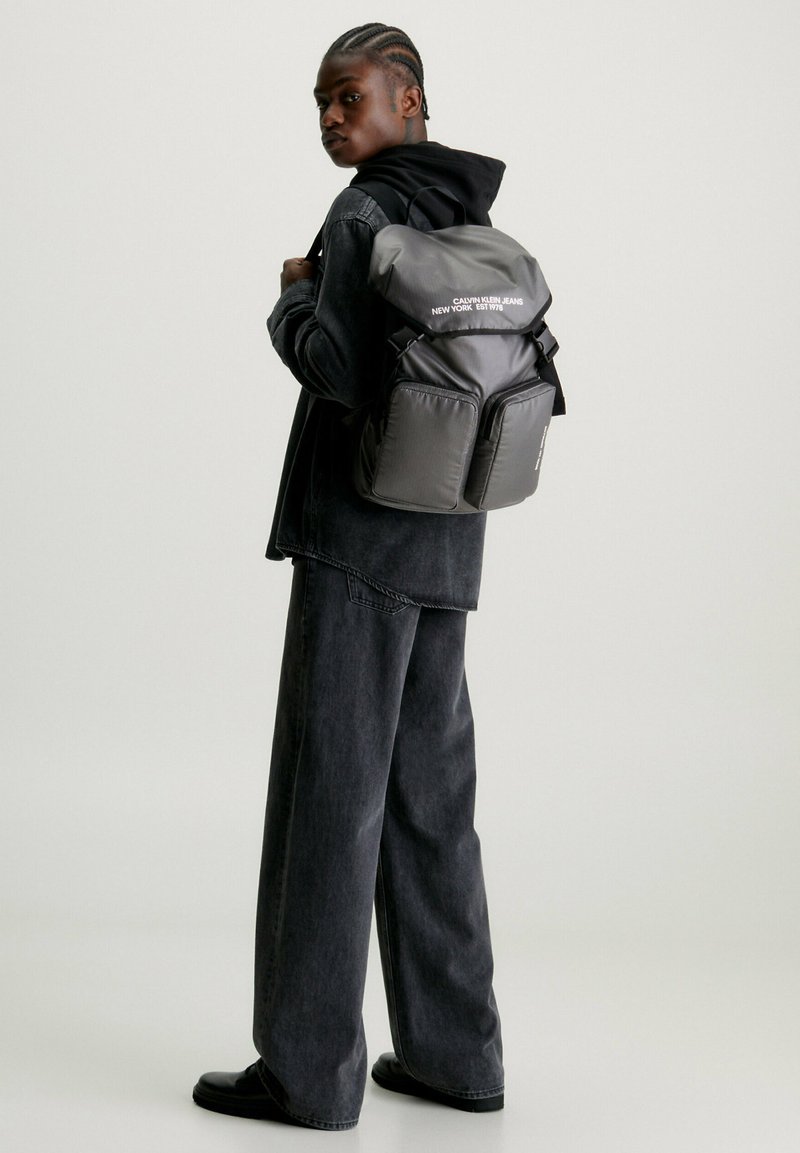 Zaino in nylon nero con apertura arrotolata, due tasche frontali e testo bianco. Il modello indossa abbigliamento scuro e si trova di fronte a uno sfondo neutro.