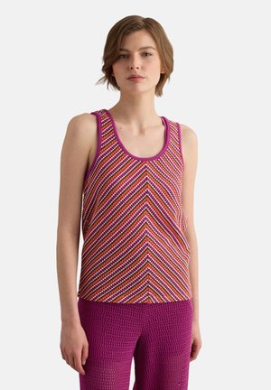 Vrouw draagt een mouwloos gestreept topje in roze, oranje en wit met paarse bies, gecombineerd met paarse gestructureerde broek.