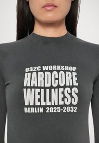 Donna che indossa una maglietta grigia scura con testo bianco che recita "032C Workshop Hardcore Wellness Berlino 2025-2032" sulla parte anteriore.