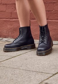 Botine negre din piele cu guler căptușit, șireturi și cusături galbene. Talpă din cauciuc groasă, texturată, cu un model de profil adânc.