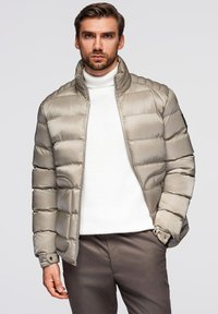 Beige pufferjakke med høj krave, vandret quilting, lynlås-lukning og sidelommer. Sammen med en hvid tekstureret sweater.