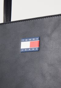 Svart teksturert håndveske med et rektangulært Tommy Jeans-logo med røde, hvite og blå aksenter som er tydelig fremstilt på den ene siden.