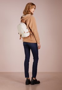Witte backpack met patronen en bruine accenten, gebogen ontwerp en verstelbare banden; gedragen met een beige trui en donkere spijkerbroek, tegen een roze achtergrond.