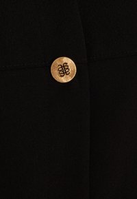 Tissu noir à surface texturée, avec un bouton rond en or embossé d'un logo stylisé. Le bouton est décoré d'un motif en forme d'éclat de soleil autour de lui.
