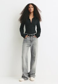 Woman with long curly hair wearing a black fitted shirt, grey wide-leg jeans with a black belt, and white sneakers, standing against a white wall.