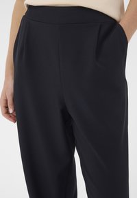 Pantalon noir taille haute avec poches latérales porté par une personne avec un haut crème, main reposant près de la poche, tissu lisse, coupe décontractée.