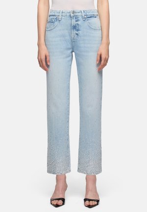 Kobieta w jasnoniebieskich dżinsach z ozdobami z kryształków skoncentrowanymi u dołu, w połączeniu z czarnymi sandałami na obcasie z odkrytymi palcami, stojąca na białym tle.