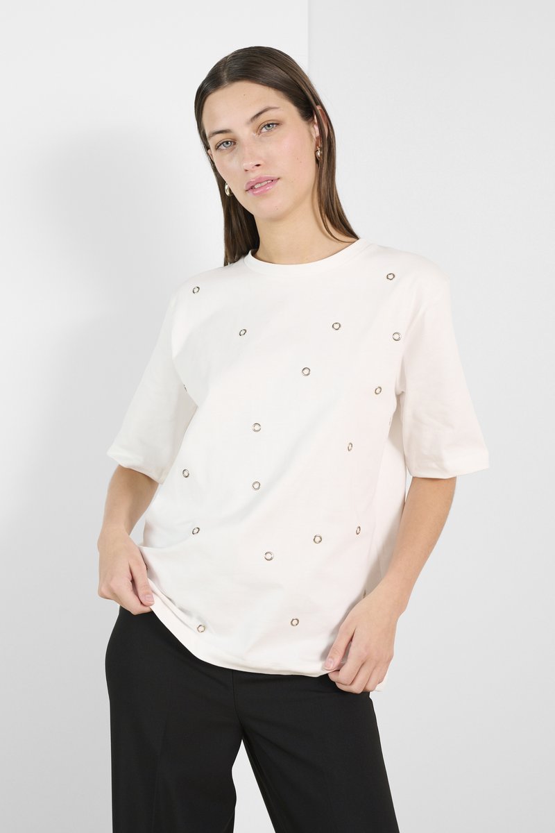 Lys beige t-shirt med korte ærmer, der har dekorative metaløjer arrangeret sporadisk over forsiden. Løs pasform.