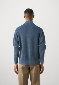 Pulover albastru cu nervuri, cu guler înalt și mâneci lungi, având o textură moale și manșete tivite, purtat cu pantaloni bej.