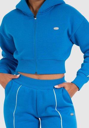Frau trägt einen knallblauen, kurzen Hoodie mit Reißverschluss und passende blaue Jogginghose mit weißen Paspeln, Hände in den Taschen.