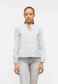 Γυναίκα με πλεξούδες, ντυμένη με λευκό πουκάμισο με μπλε λουλουδάτο σχέδιο και ανοιχτό γκρι τζιν με σκισίματα, στέκεται μπροστά σε μονόχρωμο φόντο.