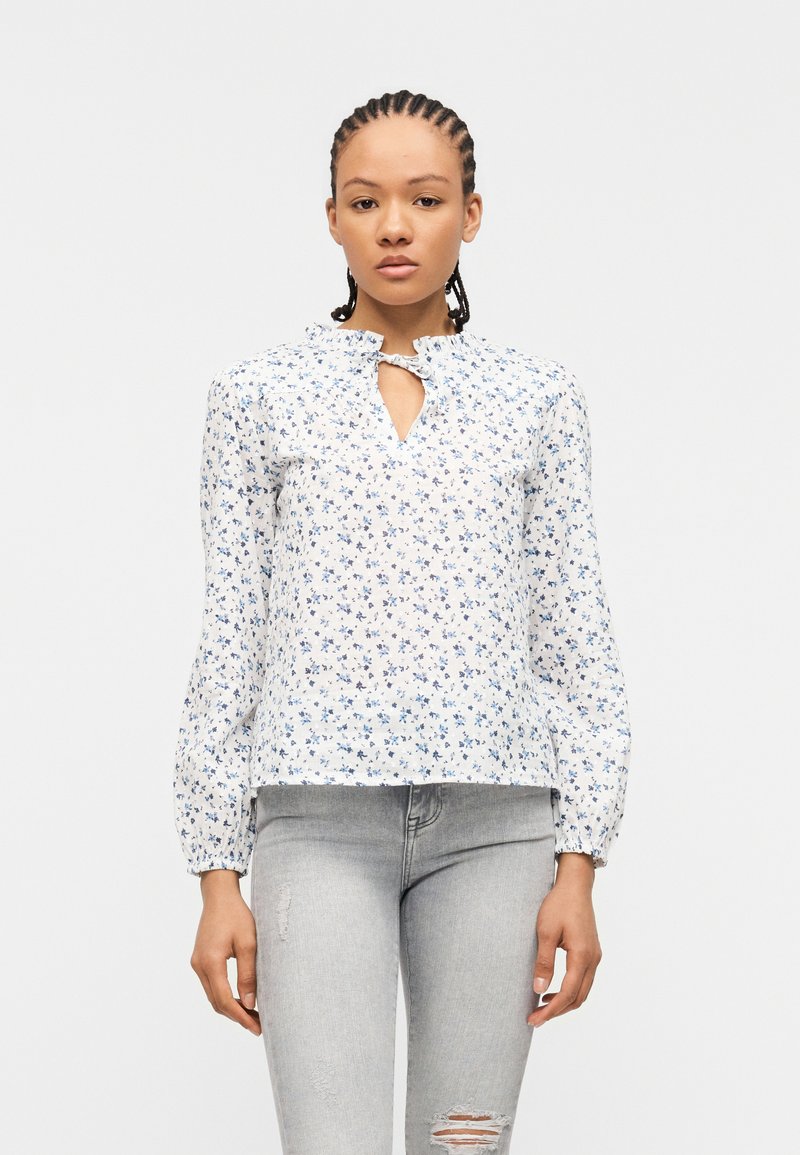 Γυναίκα με πλεξούδες, ντυμένη με λευκό πουκάμισο με μπλε λουλουδάτο σχέδιο και ανοιχτό γκρι τζιν με σκισίματα, στέκεται μπροστά σε μονόχρωμο φόντο.