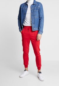 Mann in einer blauen Jeansjacke über einem weißen Rollkragenpullover, roten Jogginghosen mit weißen Streifen und weißen Sneakern, der mit den Händen in den Taschen dasteht.