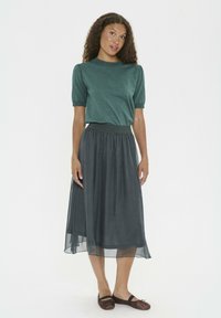 Kvinna med lockigt hår som bär en grön kortärmad topp, mörk midi kjol och bruna ballerina-skor, stående mot vit bakgrund.