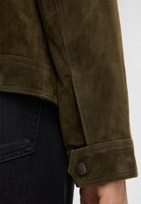 Olijfgroene suede jas, met een gemêleerde afwerking, gestikte naden en een knoopsluiting bij de manchetten. Gecombineerd met donkere denim.
