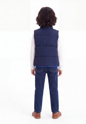 Barn med krøllet mørkt hår iført en navy polstret vest, hvid langærmet skjorte, blå jeans og brune sko, står med ryggen til.