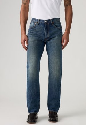 Homme portant un jean droit bleu délavé, un t-shirt blanc et des mocassins en cuir noirs, debout devant un fond clair uni.