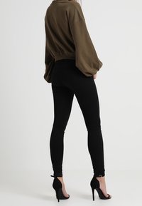Suéter oversize verde oliva con un diseño recortado, combinado con jeans ajustados negros y zapatos de tacón negros, visto desde atrás.