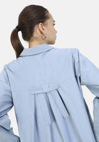 Camicia azzurro chiaro con dettaglio a pieghe sul retro, colletto e accentuazione legata. Tessuto morbido, maniche lunghe e vestibilità morbida.