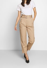 Beige high-waisted trousers with a belt featuring a metal buckle, tapered legs, and side pockets. Paired with a white T-shirt.