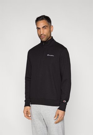 Μαύρο φούτερ με φερμουάρ κατασκευασμένο από βαμβακερό μείγμα, διαθέτει υψηλό γιακά, μανσέτες και κεντημένο λογότυπο στο στήθος. Γκρι παντελόνια φαίνονται στο κάτω μέρος.