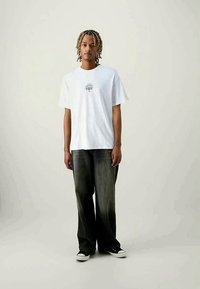 Weißes Baumwoll-T-Shirt mit kurzen Ärmeln und einem zentralen Augenmotiv. Kombiniert mit lockeren, dunkelgrauen Denim-Hosen und schwarzen Sneakern.