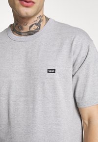 Camiseta de algodón gris con mangas cortas, que presenta un pequeño parche negro con el logo de Vans en el pecho y un cuello redondo acanalado.