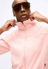 Άτομο που φοράει μεγάλα σκουρόχρωμα αθλητικά γυαλιά ηλίου ρυθμίζει το γιακά ενός ανοιχτό ροζ φλις μπουφάν με φερμουάρ και το λογότυπο της Columbia.