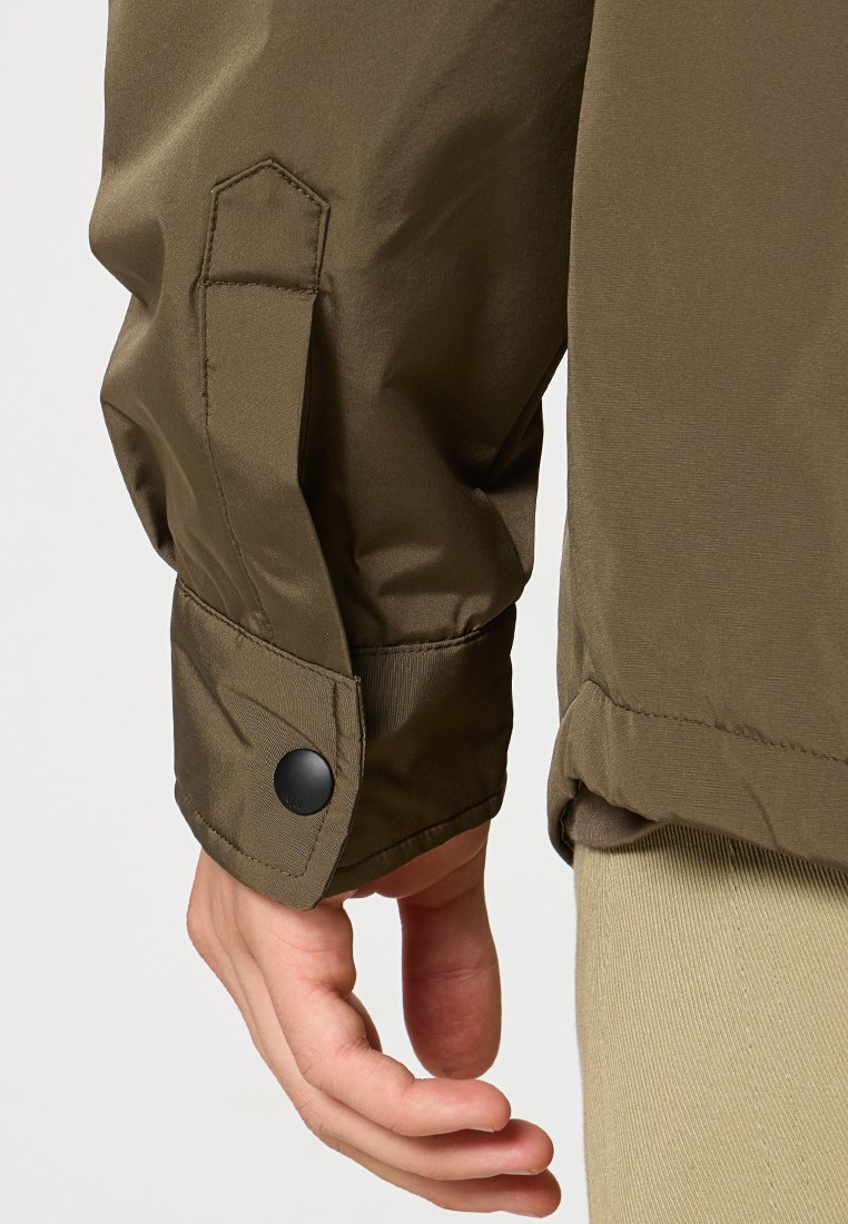 Manche de veste vert olive avec une poche à rabat, fermeture à bouton pression noir, texture lisse et détail de poignet. Main positionnée près de la veste.