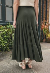 Woman wearing olive green tiered midi skirt and white ballet flats standing outdoors near plants and a small table.