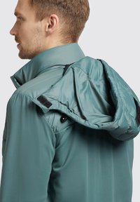Türkisfarbene isolierte Jacke mit einem Reißverschluss, hohem Kragen und einer abnehmbaren Kapuze, die mit Klettverschluss und Druckknöpfen gesichert ist. Glatte, weiche Textur.